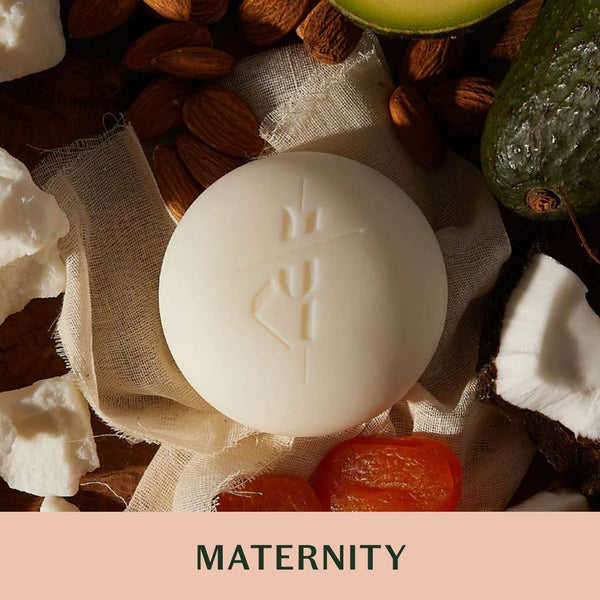 White soap bar with branding surrounded by natural ingredients on a pink background, labeled 'MATERNITY'.