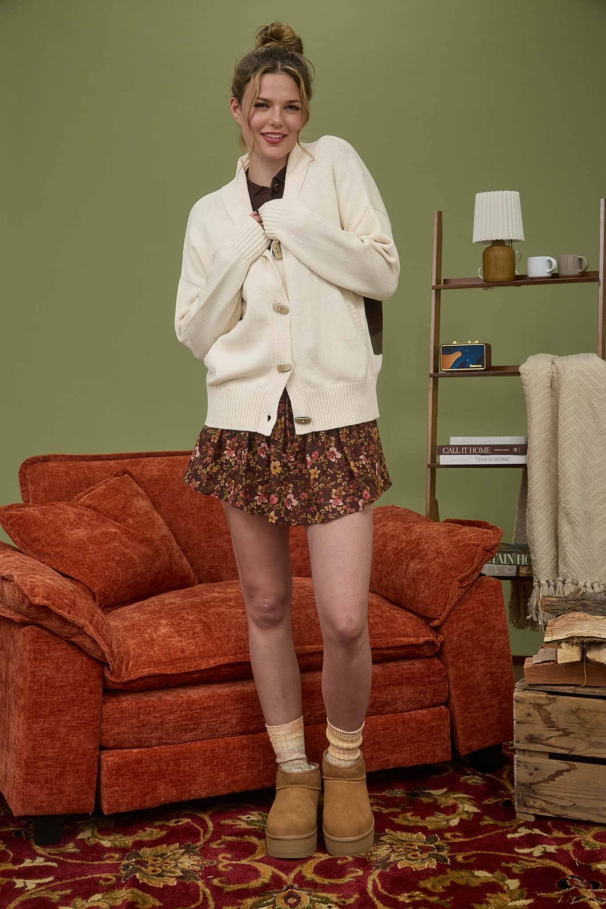 Woman in a white cardigan and floral skirt standing in a living room with a red couch and green wall.