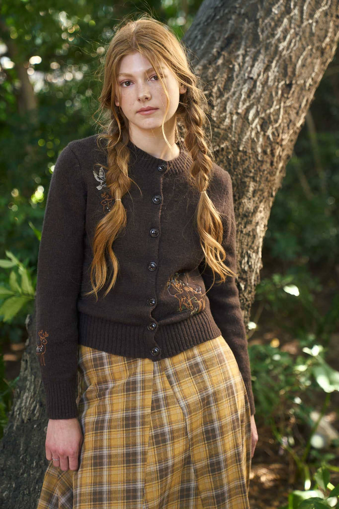 Woman in a brown cardigan and plaid skirt standing in a forest